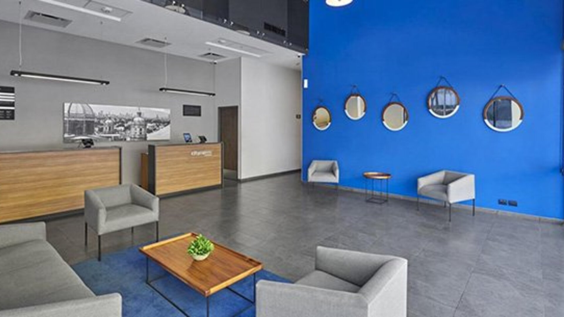 Hotel reception with wood-fronted desks, grey sofas, round wall mirrors and a bright blue accent wall at City Express by Marriott CDMX La Villa.