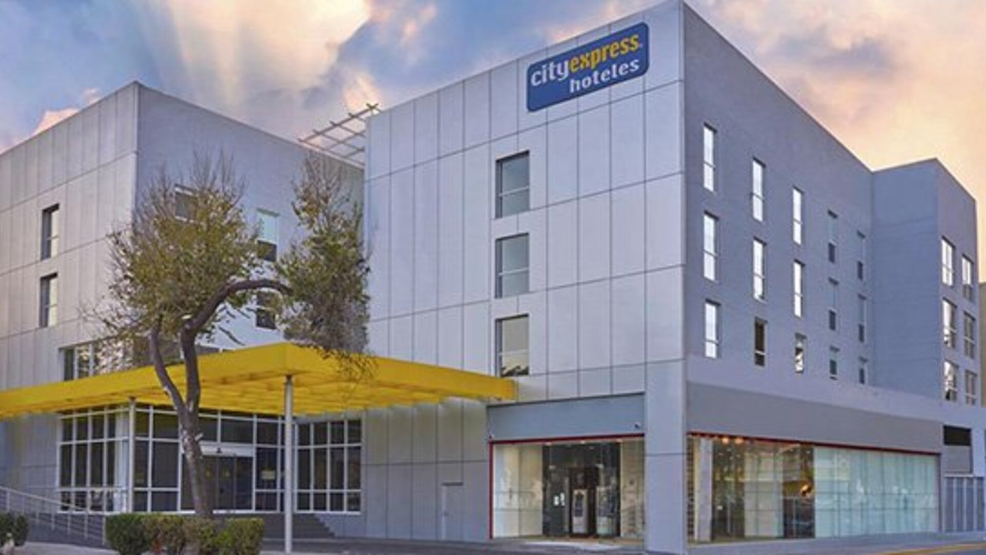 Modern hotel exterior with silver panelled walls, large windows and a yellow entrance canopy at City Express by Marriott CDMX La Villa.