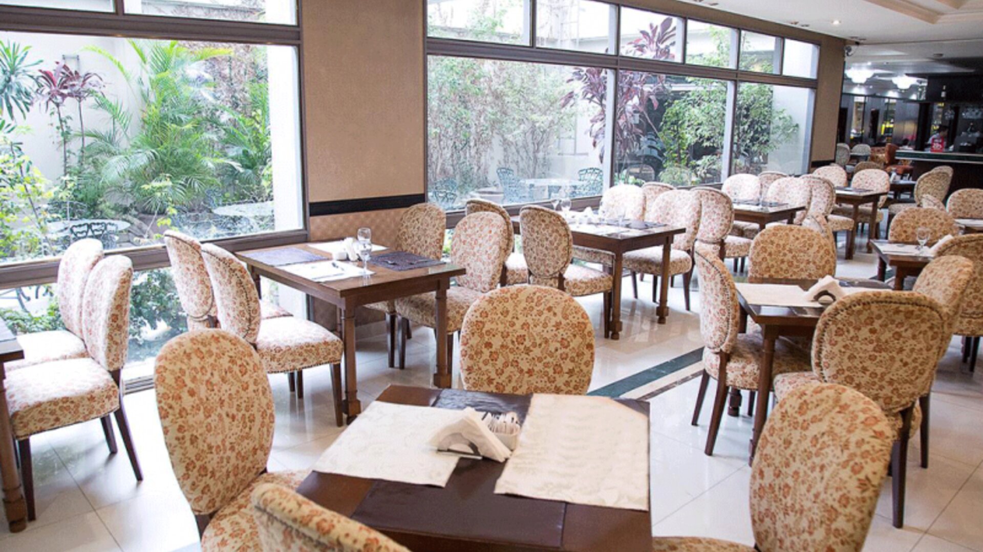 Dining room with patterned upholstered chairs, wooden tables, white tiled floors and large garden-facing windows at HR Esmeralda Luxor.