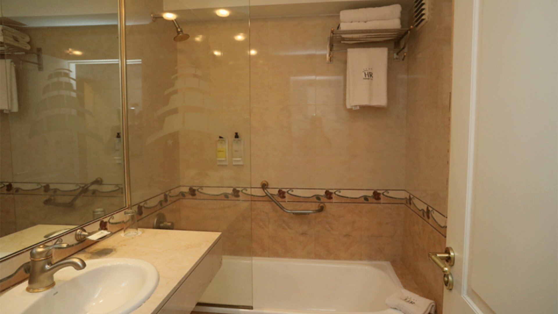 Bathroom with beige tiled walls, glass shower screen, bathtub, white vanity sink and grab rails at HR Esmeralda Luxor.