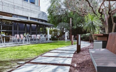 Outdoor garden walkway with a wooden bench, trees, green lawn and restaurant terrace at Hotel Best Western PLUS Alfa Aeropuerto.