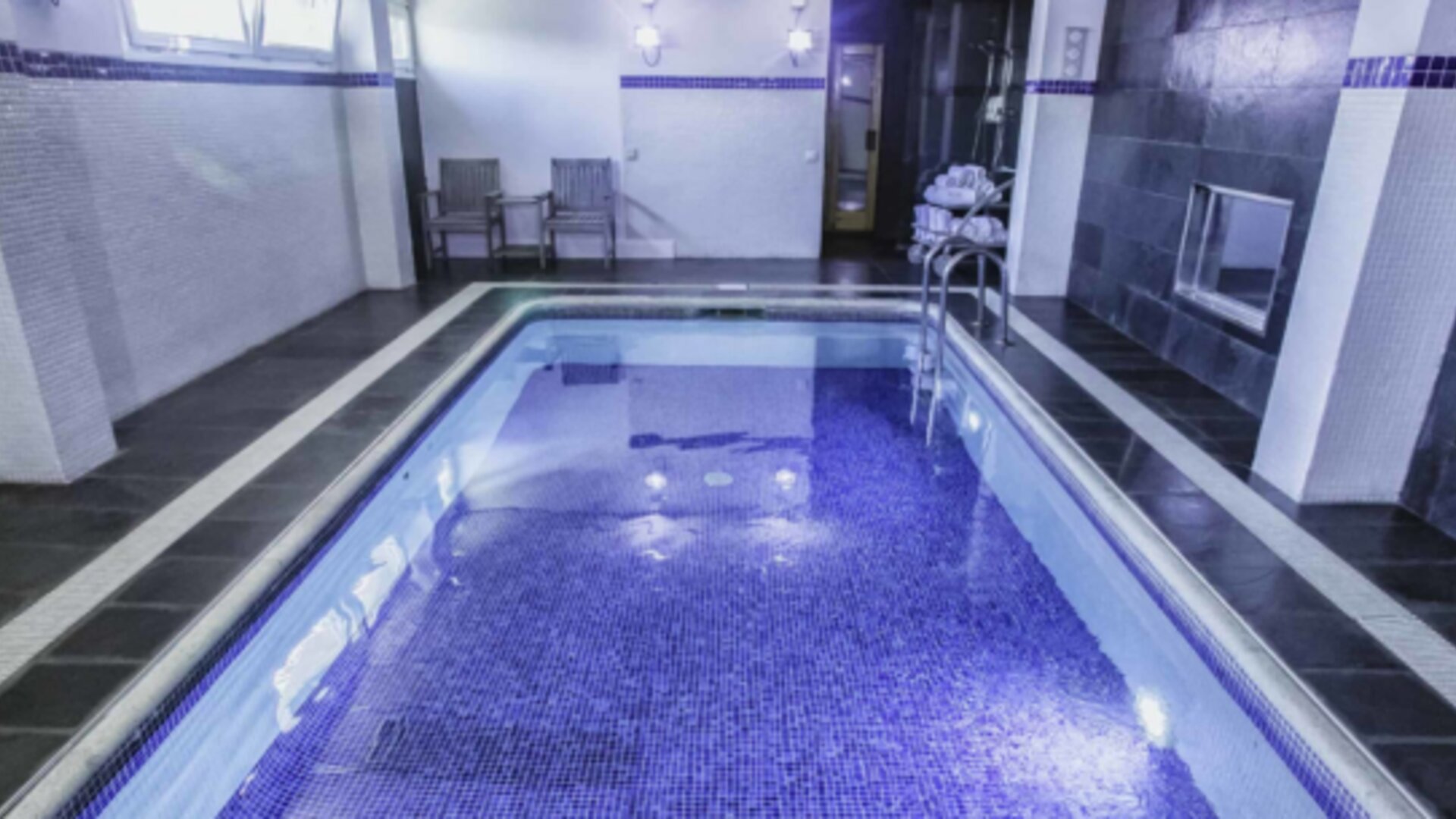 Indoor swimming pool with blue mosaic tiles, dark stone flooring, white walls and a metal handrail at Hotel Best Western PLUS Alfa Aeropuerto.