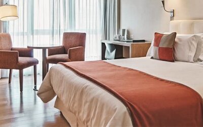 Guest room with double bed, rust-coloured bed runner and cushions, two armchairs and large curtained windows at Hotel Regente Palace.