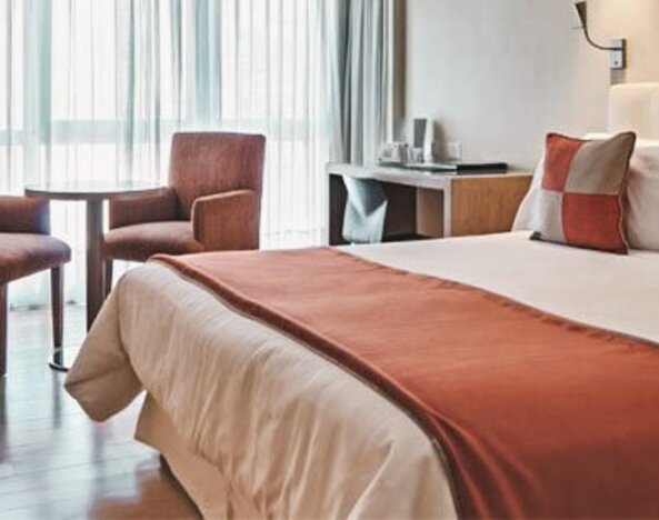 Guest room with double bed, rust-coloured bed runner and cushions, two armchairs and large curtained windows at Hotel Regente Palace.