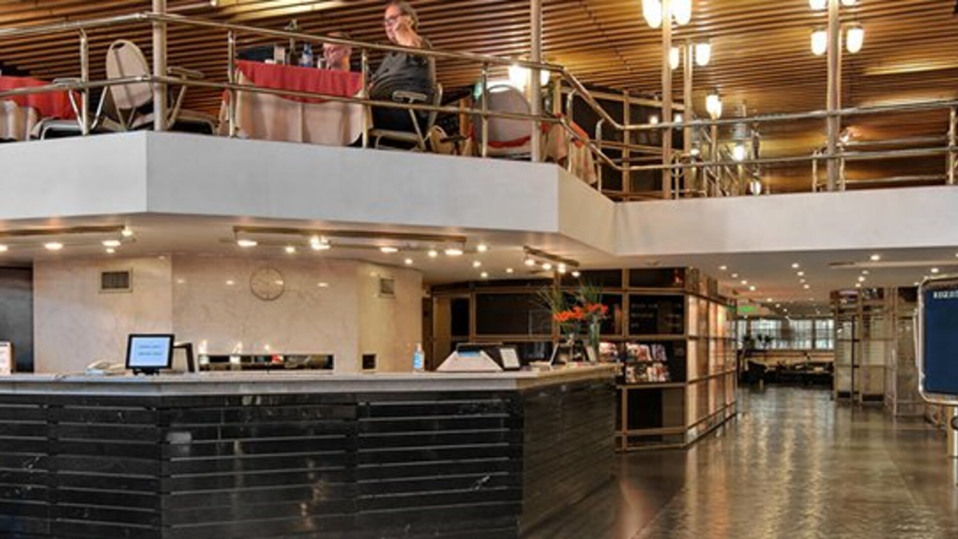 Hotel reception with black front desk, upper-level dining area, metal railings and wooden slat ceiling at Hotel Regente Palace.