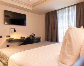 Guest room with white bedding, wall-mounted television, desk, dark curtains and bedside lighting at Hotel Silken Saaj Las Palmas.