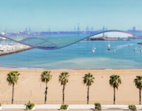 Coastal view of a sandy beach, palm-lined promenade, blue sea and distant harbour beneath a clear sky at Hotel Silken Saaj Las Palmas.