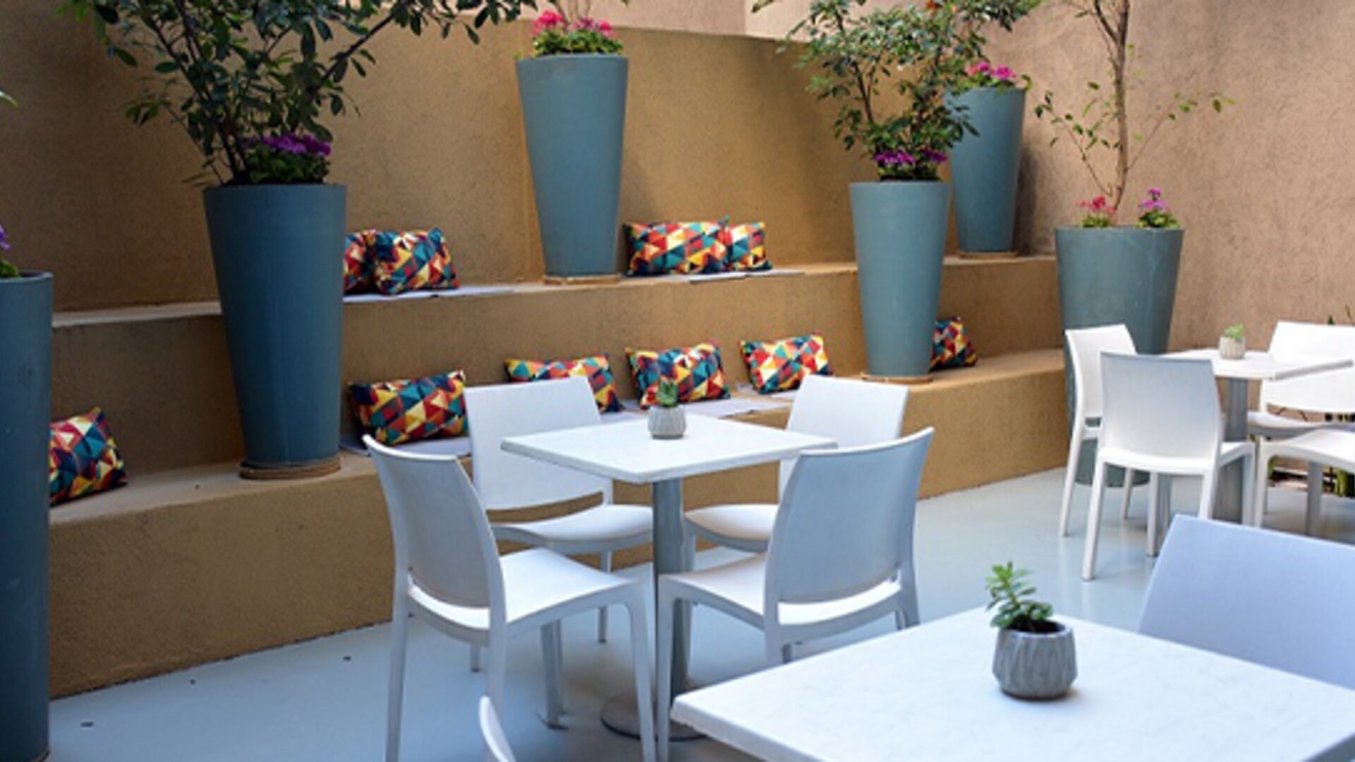 Outdoor dining patio with white tables and chairs, tall blue planters and colourful cushions at Ian Hotel.
