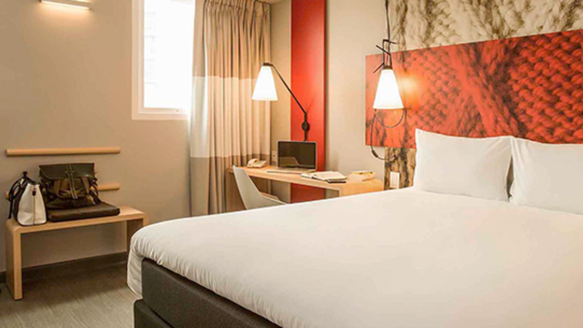 Guest room with a large white bed, red accent headboard, bedside lamps, desk and curtained windows at Ibis Lima Reducto Miraflores.