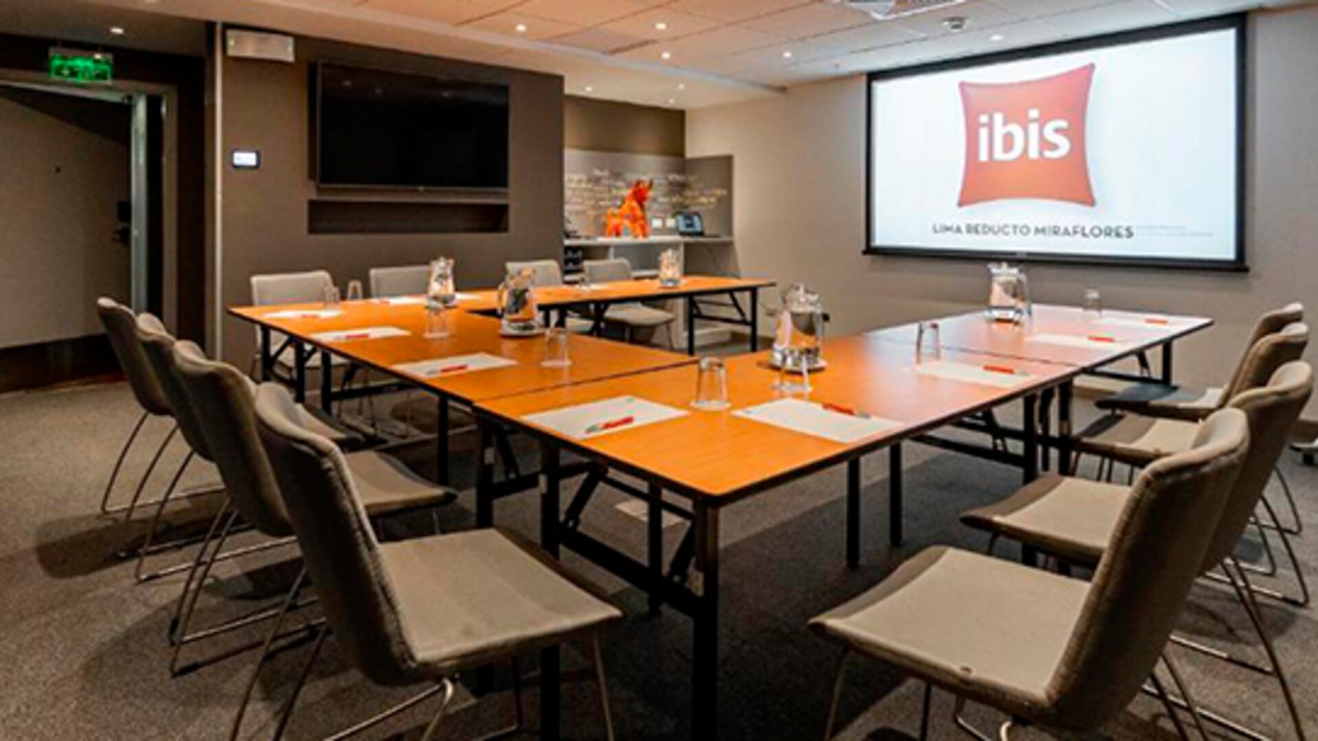 Meeting room with conference tables, grey chairs, water glasses and a presentation screen at Ibis Lima Reducto Miraflores.