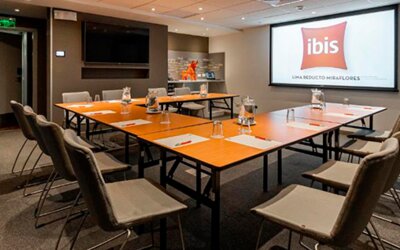 Meeting room with conference tables, grey chairs, water glasses and a presentation screen at Ibis Lima Reducto Miraflores.