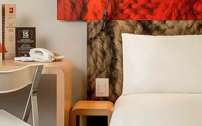 Guest bed with white bedding and pillows beside a wooden desk, chair and bedside table at Ibis Lima Reducto Miraflores.