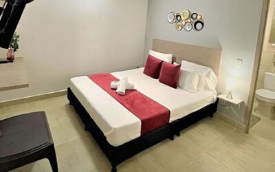 Guest room with double bed, red runner and cushions, wall-mounted television and ensuite bathroom visible through the doorway at 12 Nights Hotel.
