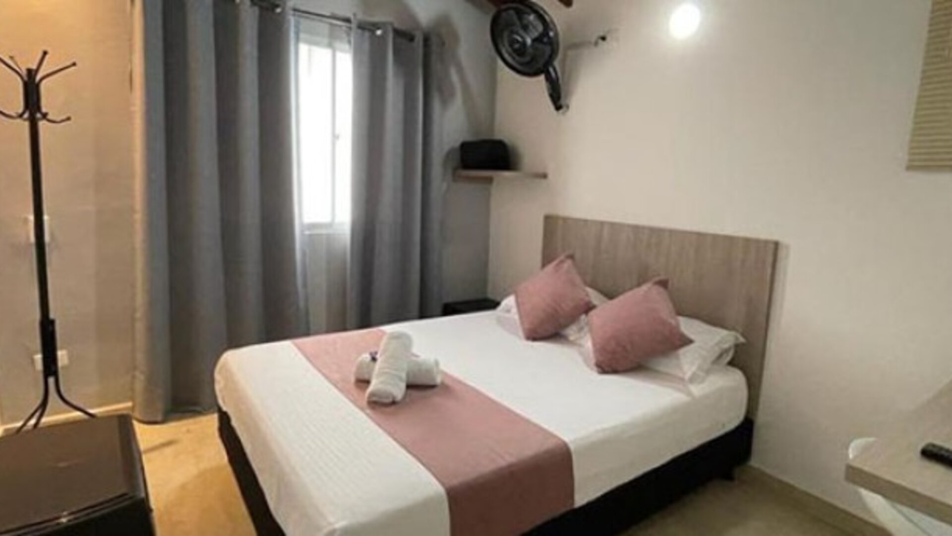 Guest room with double bed, pink accent cushions and runner, grey curtains, wall fan and wooden desk at 12 Nights Hotel.