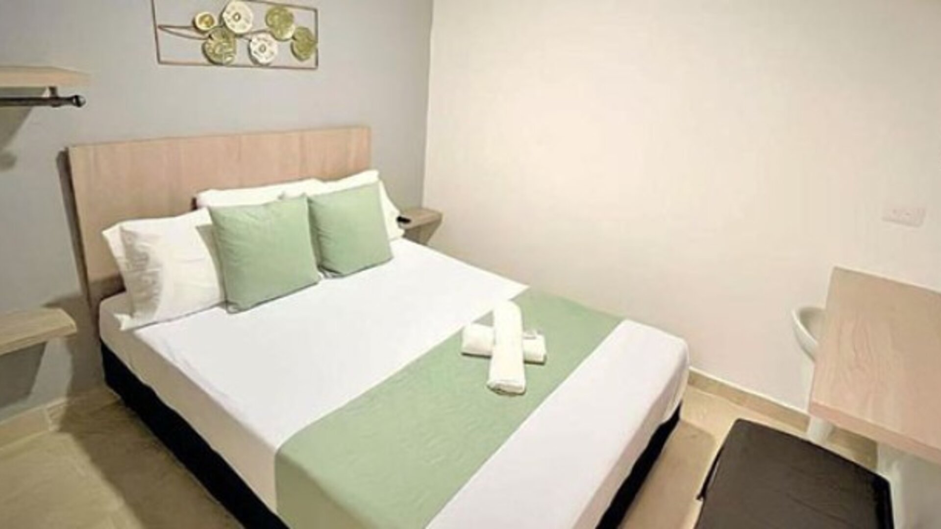 Guest room with double bed, green accent cushions, matching bed runner, wall-mounted television and wooden desk at 12 Nights Hotel.