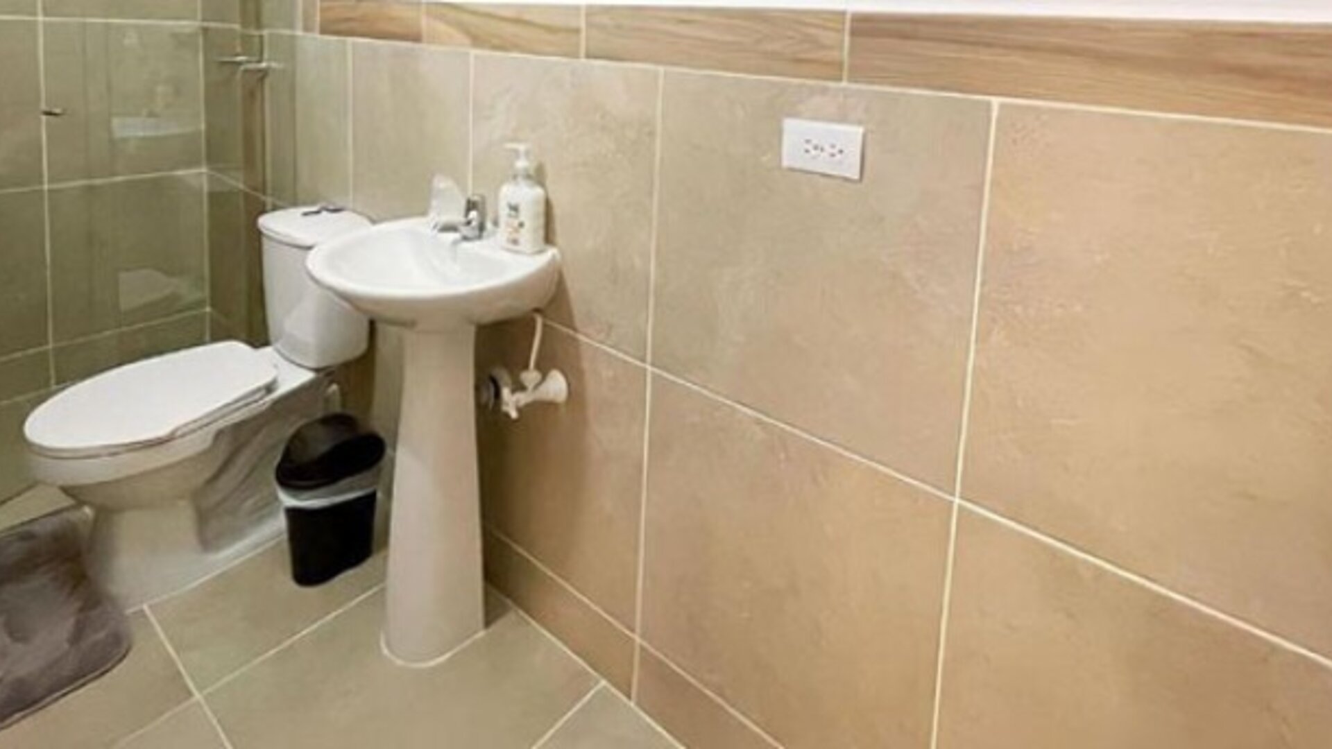 Bathroom with beige tiled walls, glass shower enclosure, pedestal sink, toilet and soap dispenser at 12 Nights Hotel.