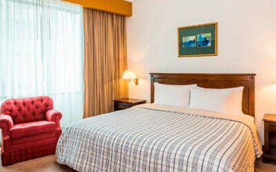 Guest room with double bed, striped bedding, red armchair, bedside lamps and a large curtained window at Hospedium Princess Hotel Panamá.