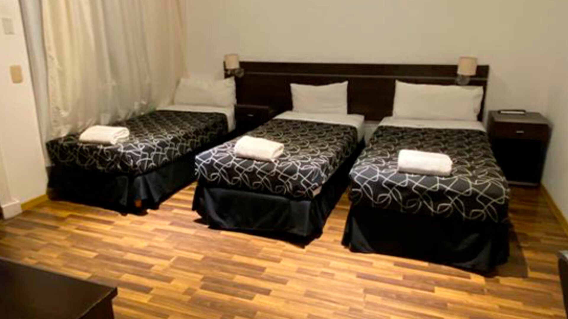 Guest room with three single beds, black patterned bedding, white towels, bedside lights and wooden flooring at Bagu Buenos Aires.