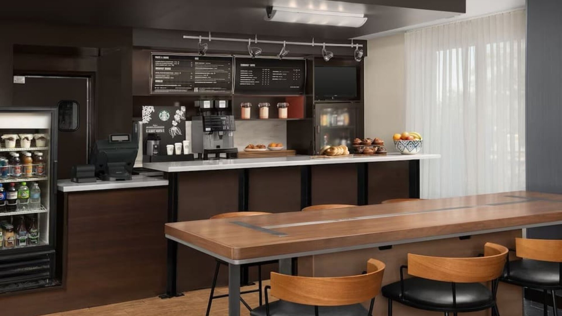 Refreshment station with coffee machines, refrigerated drinks, pastries, fruit and a long communal wooden table at Courtyard By Marriott Dothan.