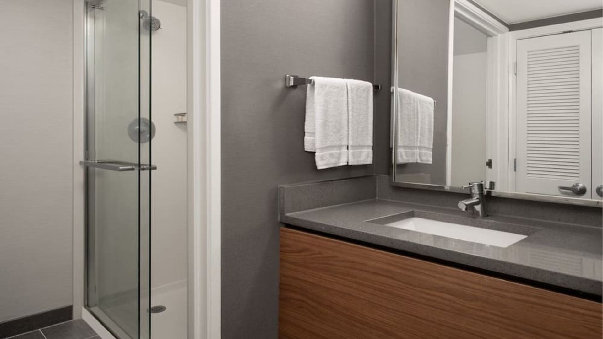 Bathroom with glass shower enclosure, wall-mounted showerhead, gray vanity countertop, inset sink and folded white towels at Courtyard By Marriott Dothan.