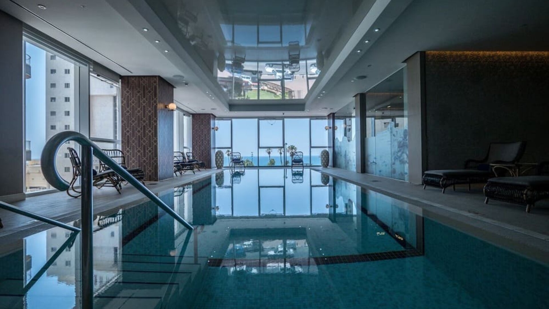 Indoor swimming pool with floor-to-ceiling windows, water views and dark wicker loungers at David Tower Hotel Netanya.