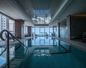 Indoor swimming pool with floor-to-ceiling windows, water views and dark wicker loungers at David Tower Hotel Netanya.