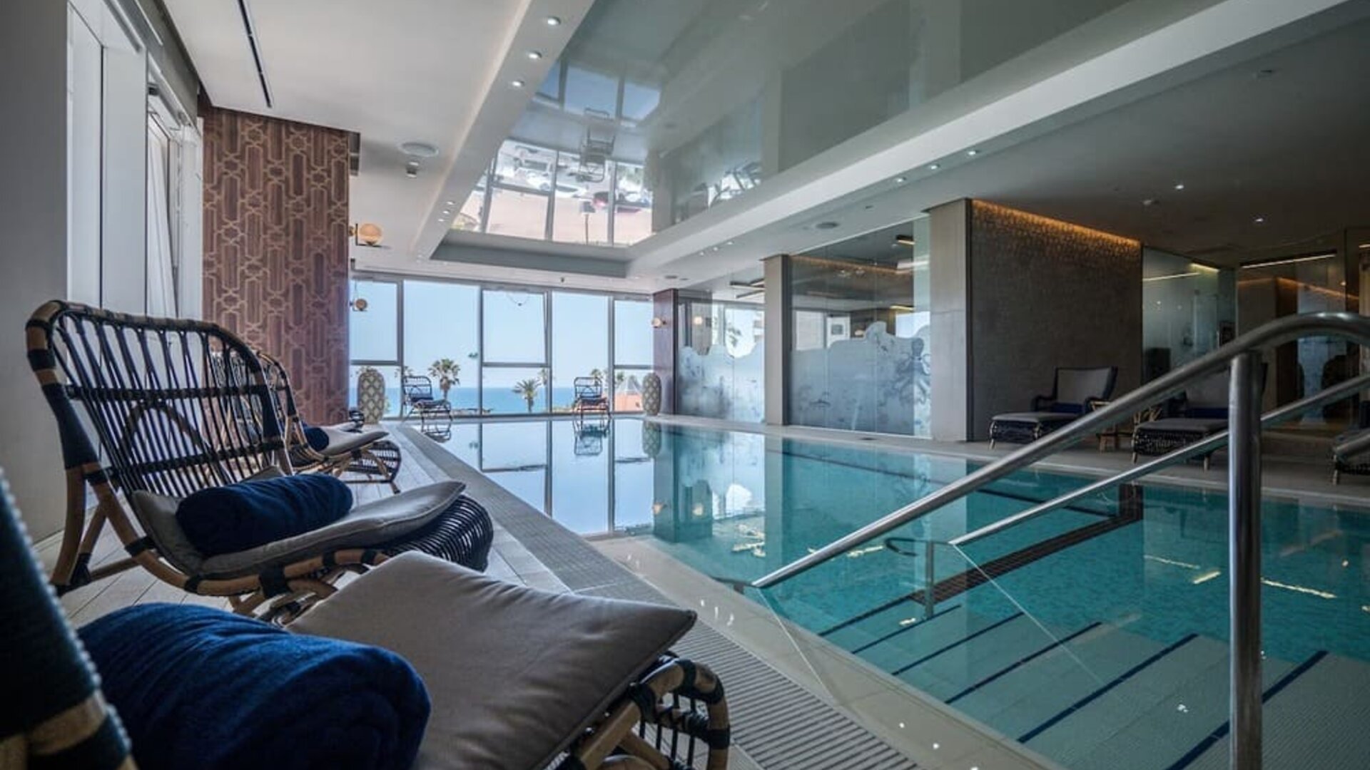 Indoor pool viewed beside wicker loungers, with glass walls, steps and sea views through the windows at David Tower Hotel Netanya.