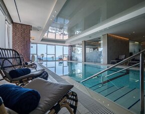 Indoor pool viewed beside wicker loungers, with glass walls, steps and sea views through the windows at David Tower Hotel Netanya.