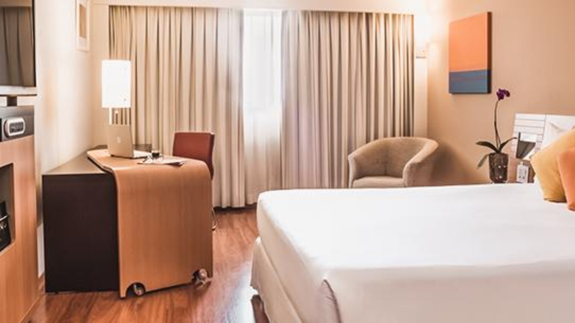 Guest room with king bed, orange accent cushions, wooden desk, armchair, television and floor-to-ceiling curtains at Novotel Lima San Isidro.