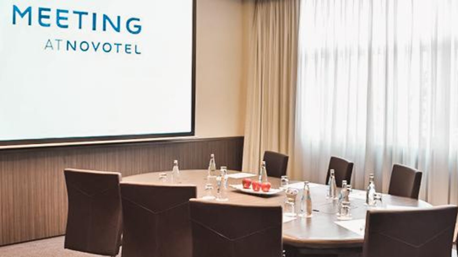 Meeting room with a conference table, brown chairs, presentation screen and large windows with sheer curtains at Novotel Lima San Isidro.