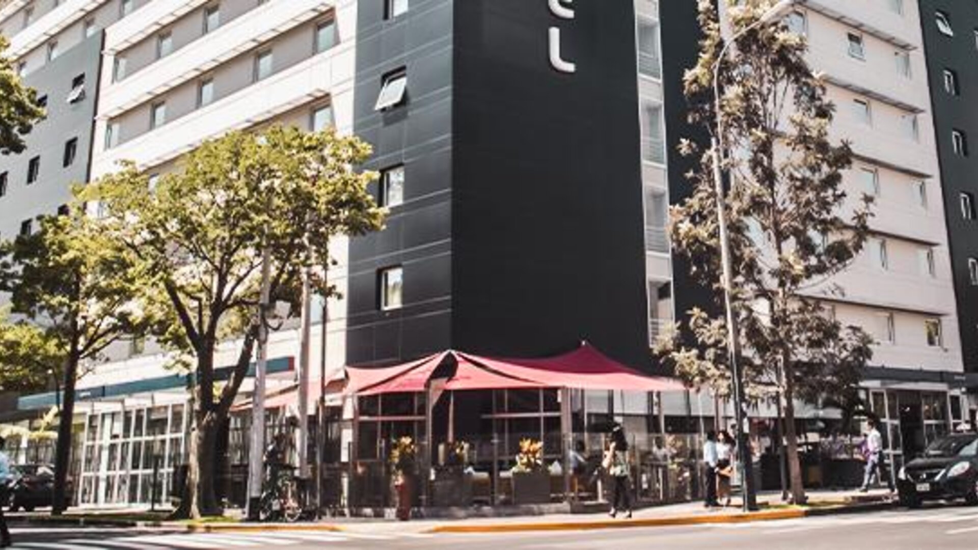 Exterior view of the modern hotel building with street-level windows, red entrance awnings, trees and sidewalk seating at Novotel Lima San Isidro.