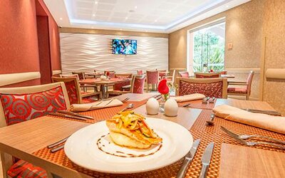 Restaurant dining area with set tables, red patterned chairs, plated food and wall-mounted television at Apartamentos Plaza Suites.