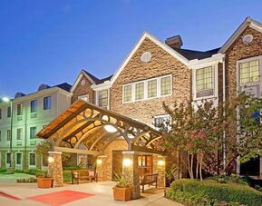 Sonesta ES Suites Dallas Las Colinas’ exterior has a covered entrance, benches, and greenery including potted plants, well-kept bushes, and trees.