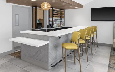 Hotel bar at Hilton Garden Inn Queens/JFK Airport with white counter, yellow stools, and bottle display.