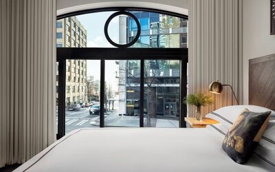 Bedroom with large window showing city street view, white bedding, and neutral decor at Kimpton Palladian Hotel.