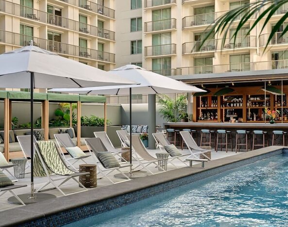 Outdoor pool with sun loungers, umbrellas, tiled deck, and mural wall surrounded by greenery at Romer House Waikiki.