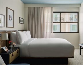 Queen bed with woven headboard beside window, soft curtains and framed wall art in a cozy layout at Romer House Waikiki.
