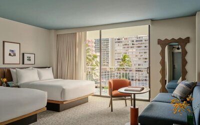 Guest room with beds near large window, balcony view of buildings, and seating area with chair and table at Romer House Waikiki.