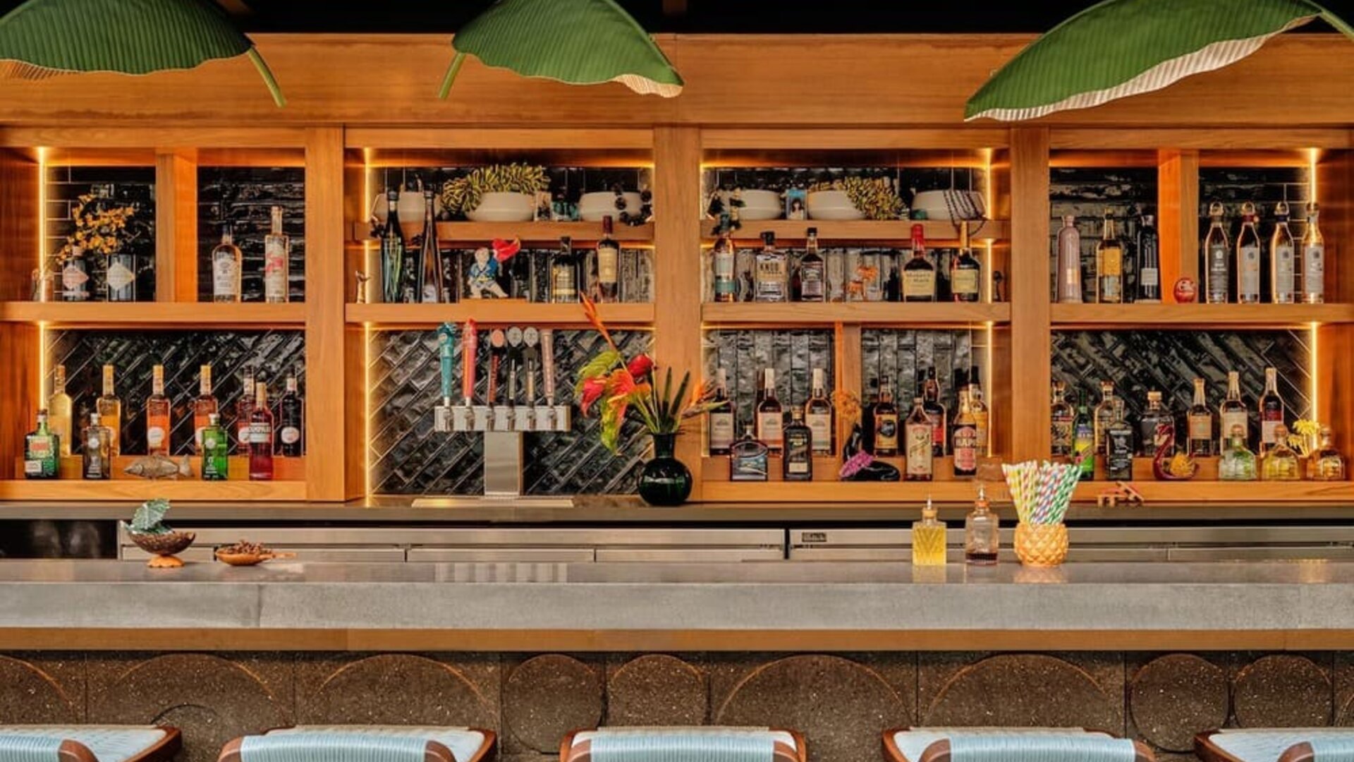 Bar with wooden shelving, green leaf pendant lights, and a wide counter lined with stools at Romer House Waikiki.