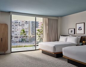 Two beds with white linens, balcony access, city view, and seating area with sofa and table at Romer House Waikiki.