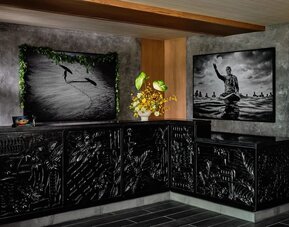 Reception desk with dark carved front panels, floral arrangement, and framed black-and-white artwork behind at Romer House Waikiki.