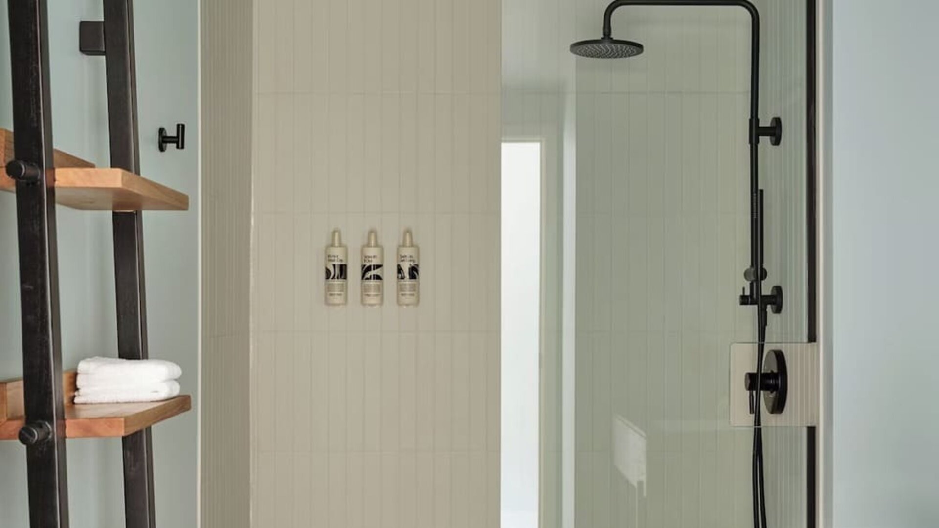 Bathroom with walk-in glass shower, black fixtures, white tile walls, and wooden shelving with towels at Romer House Waikiki.