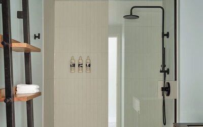 Bathroom with walk-in glass shower, black fixtures, white tile walls, and wooden shelving with towels at Romer House Waikiki.