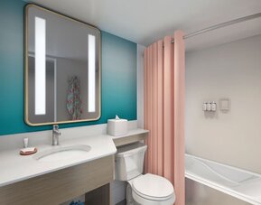 Bathroom with vanity, illuminated mirror, bathtub with pink curtain, and teal accent wall at The Twin Fin Hotel.