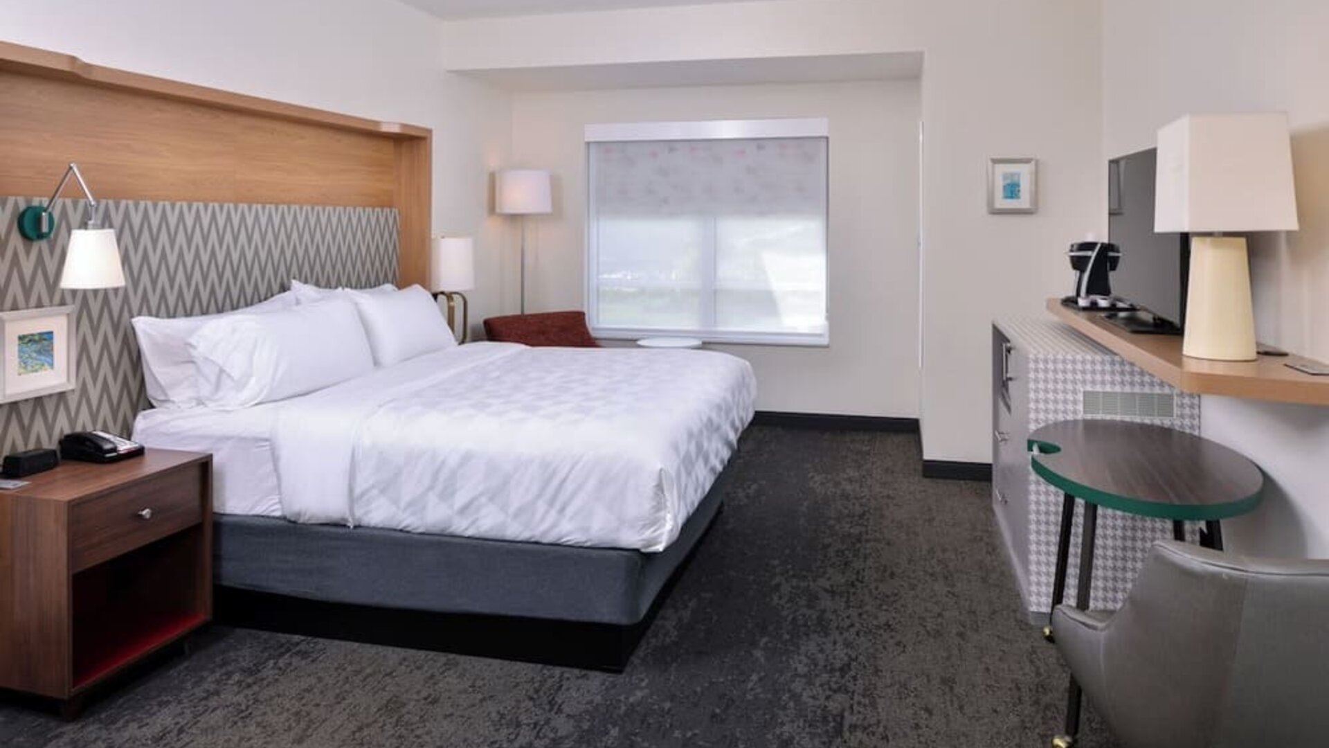 Spacious room with work desk, chair, TV stand, and modern lighting at Holiday Inn & Suites Farmington Hills - Detroit NW By IHG.