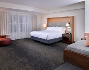 Large suite with king bed, sofa seating, and wide window with sheer curtains at Holiday Inn & Suites Farmington Hills - Detroit NW By IHG.