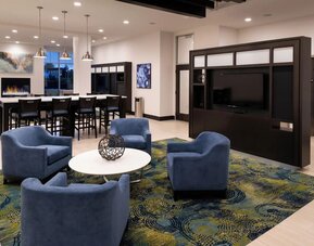 Lounge area with chairs, TV wall, communal table, and fireplace at Holiday Inn & Suites Farmington Hills - Detroit NW By IHG.