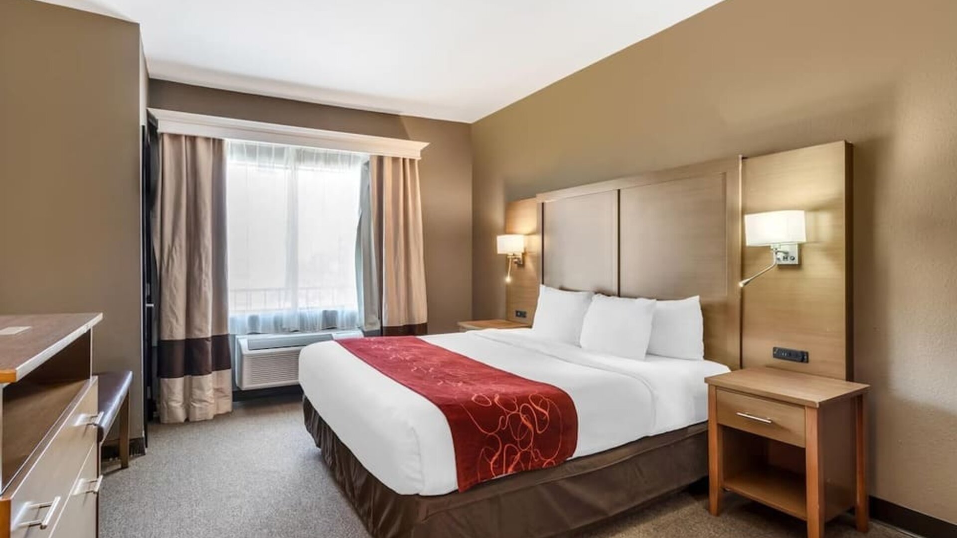 King bedroom with bedside tables, lamps, and window with curtains and air conditioning unit at Comfort Suites Bush Intercontinental Airport.