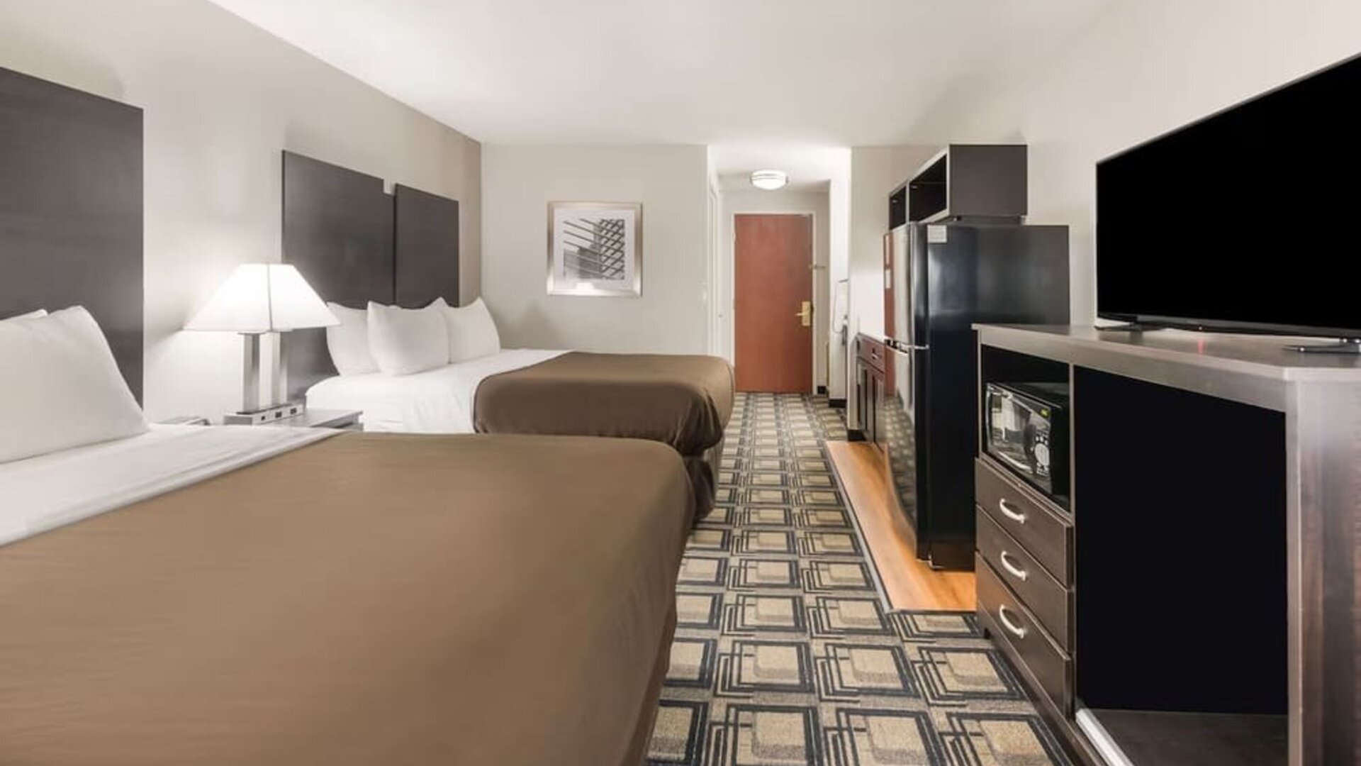 Room with two beds, dresser, TV, microwave, mini fridge, and patterned flooring at Suburban Studios Mason Hwy-42.