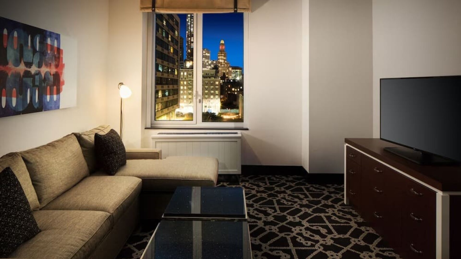 Living area with sofa, coffee table, television, and city view through window at Hilton Brooklyn New York.